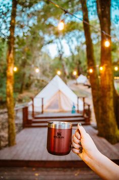 a person holding a coffee cup in front of a wooden deck with lights strung from the trees