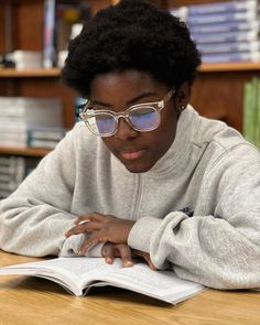 a woman in glasses reading a book at a table with bookshelves behind her