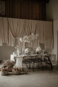 the table is set with white flowers and vases in front of curtains that are drapes