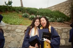 two women in graduation gowns holding books and posing for the camera with their arms around each other