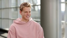 a man in a pink hoodie smiles at the camera while standing next to a building