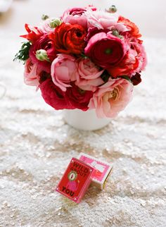 a white vase filled with red and pink flowers next to a matchbox on a table