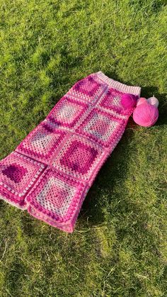 a pink crocheted blanket laying on the grass next to a ball of yarn