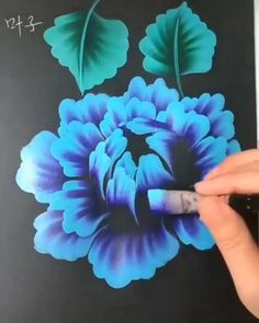 a person is painting a flower with blue and green flowers on black paper that has leaves in the center