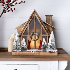 a christmas scene with a lit candle and a small house on a table in front of a white painted wall