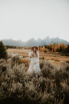 a woman in a white dress and hat walking through a field with mountains in the background