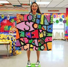 a woman holding up a large painting in a room filled with children's artwork