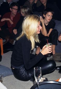 a woman sitting on the floor with a drink in her hand and people behind her