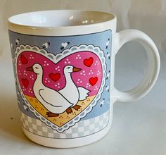 a white coffee mug with two geese on it's side and hearts in the background