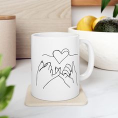 two hands making a heart with their fingers on a white coffee mug next to some fruit