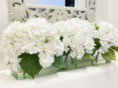 white flowers are in a glass vase on a mantle with a mirror behind it and a decorative frame