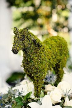 a moss covered horse standing on top of a white table cloth next to flowers and greenery