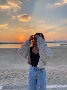 a woman is standing on the beach with her hands to her face as the sun sets