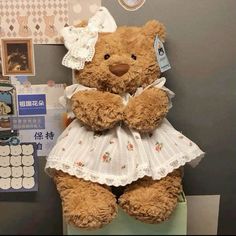 a brown teddy bear wearing a white dress sitting on top of a desk next to a clock