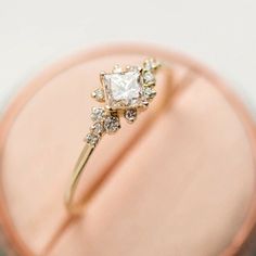 an engagement ring with a princess cut diamond surrounded by small white and clear stones on a pink box