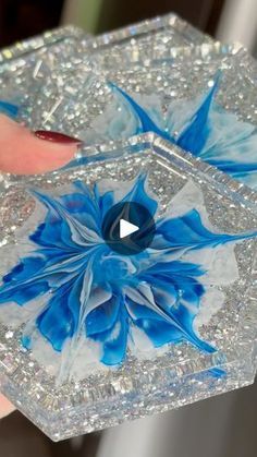 a hand holding a blue and white flower in a glass box with glitters on it