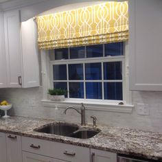 a kitchen sink under a window with yellow curtains on the windowsill and white cabinets