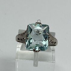 Set Securely In The Center Of The Ring Is A Large Emerald Cut Aquamarine Gemstone (*I Am Unsure If This Stone Is Natural Or Created) It Has A Beautiful Medium Icy Blue Color With Very Nice Sparkle. **There Is A Chip In One Corner Of The Stone-See Photos!** The Aquamarine Measures 11 X 9mm And Is Set With Four Double Claw Prongs. The Prongs Are Set At North, South, East And West Instead Of The Traditional Four Corners. The Setting Has Delicate Cutwork All Underneath The Center Gem And Down The Band. There Is A Milgrain Detailing In The Scroll Along With Four Little Flowers. The Band Has A Shiny High Polished Finish That Would Be Easy To Size Up Or Down (Locally). The Setting Is Solid White Go Elegant Emerald Cut Blue Gemstones, Elegant Blue Emerald Cut Gemstones, Classic Sapphire Gemstones With Accent Stones, Elegant Solitaire Topaz Ring For Formal Occasions, Elegant Formal Solitaire Topaz Ring, Classic Blue Topaz Ring For Formal Occasions, Classic Formal Blue Topaz Ring, Classic Aquamarine Gemstone Jewelry, Classic Blue Topaz Diamond Cut Ring