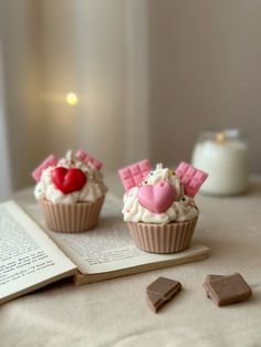 two cupcakes with hearts and marshmallows sitting on top of an open book