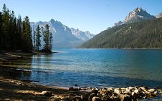 a lake surrounded by mountains and trees with rocks in the foreground on a sunny day