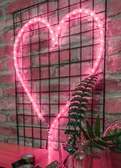 a pink heart shaped neon sign sitting on top of a table next to a potted plant