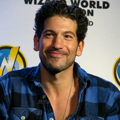 a man with curly hair smiles at the camera while wearing a blue shirt and black checkered shirt