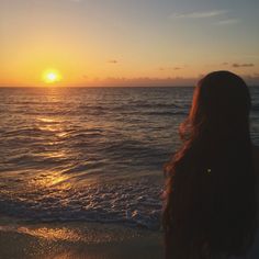 a woman standing on top of a beach next to the ocean at sunset with her hair blowing in the wind