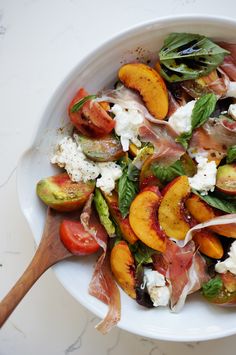 a salad with peaches, cheese and spinach in a white bowl next to a wooden spoon