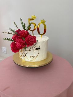 a white cake with red roses and gold numbers on it sitting on a pink table