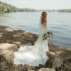 a woman in a wedding dress standing on rocks near the water holding a bouquet and looking off into the distance