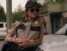 a woman police officer standing in front of her patrol car wearing sunglasses and a hat