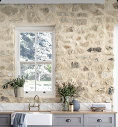 a kitchen with stone wall and white sink in the center, windows above it are potted plants