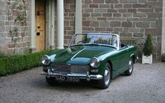 an old fashioned green car parked in front of a brick building next to a hedge