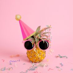 a pineapple wearing sunglasses and a party hat with confetti around it on a pink background