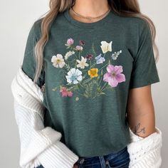 Make a statement with our pressed flower shirt! The beautiful wildflower graphic on this shirt is sure to turn heads and add a touch of nature to any outfit. The graphic is designed with vibrant colors and intricate details that will make you feel like you're wearing a work of art.  Not only is our Flower Shirt stylish, but it's also made from high-quality materials that are soft and comfortable to wear. The shirt is easy to care for, so you can wear it again and again without worrying about it losing its shape or color. This shirt makes a perfect gift for any fashion-forward woman in your life. Whether it's for a birthday, anniversary, or just as a surprise, our Flower Shirt is sure to be a hit. It's versatile enough to wear to work or out on the town, making it a wardrobe staple that she Green Graphic Tee With Plant Print, Green Graphic Tee With Plants Print, Botanical Flower Graphic Print T-shirt, Botanical Style Flower Graphic T-shirt, Spring Botanical T-shirt With Floral Design, Green Botanical Short Sleeve Top, Spring Botanical Flower T-shirt, Botanical Screen Print Tops For Spring, Botanical Style Screen Print Tops For Spring