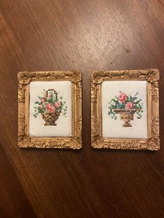 two framed cross - stitch pictures on a wooden table, one with flowers in it