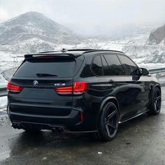 the rear end of a black bmw suv parked on a road in front of snow covered mountains