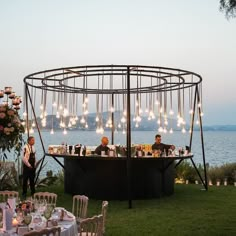 two people are sitting at an outdoor bar with lights hanging from the roof and chairs around it