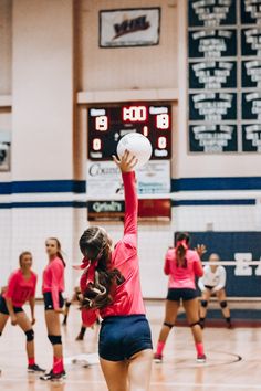 the girls are playing volleyball together on the court in the gym, and one girl is reaching up to hit the ball with her hand