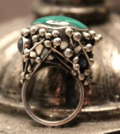 Add a unique touch to your jewelry collection with this stunning handmade abstract ring. The ring features a beautiful cabochon-shaped malachite gemstone, set in sterling silver. The green-colored ring is a perfect accessory for any occasion. The ring is crafted in the United States and has a ring size of 8. The secondary stone, sapphire, adds to the uniqueness of the ring. The malachite cabochon is the main attraction, but the sapphire stone adds an extra sparkle to the piece. The ring is perfect for those who appreciate fine jewelry and want to add a unique touch to their collection. Green Gemstone Cabochons Fine Jewelry, Green Oval Cabochon Dome Ring, Formal Malachite Gemstone Rings, Modernist Round Cabochon Jewelry, Elegant Green Chrysocolla Jewelry, Green Malachite Rings With Polished Finish, Elegant Malachite Gemstone Ring, Modernist Oval Cabochon Rings, Unique Green Cabochon Ring