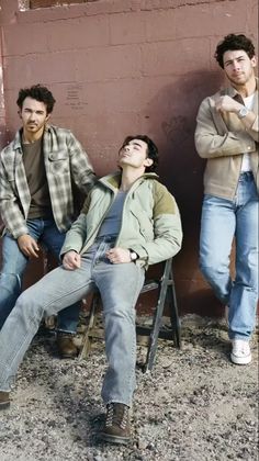 three young men sitting next to each other in front of a red wall and one man laying on a folding chair