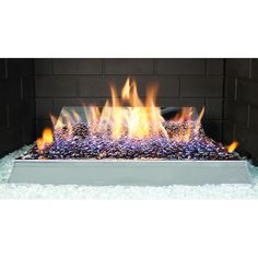 an outdoor fire pit with flames and rocks on the bottom, in front of a brick wall