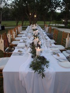 a long table is set with white linens and place settings for an outdoor dinner