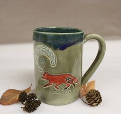 a green mug with a red fox on it next to some pine cones and leaves