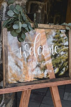 a welcome sign with greenery on it in front of a stone wall and wooden easel