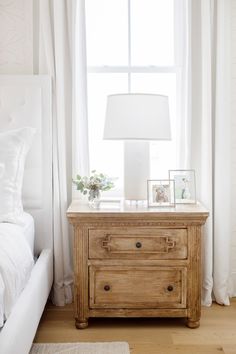 a bedroom with a bed, nightstand and window in the corner that has white curtains