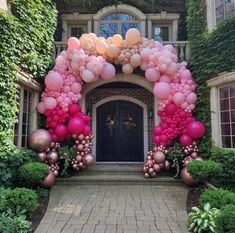 an archway decorated with pink and gold balloons in front of a brick building surrounded by greenery