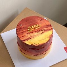 a round cake sitting on top of a white paper covered box with gold lettering and frosting