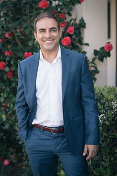 a man in a blue suit and white shirt smiles at the camera with red roses behind him