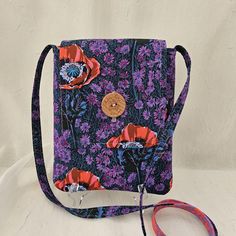 When you need a smaller bag to carry just the essentials, select an Autumn Rain Gallery crossbody bag. This stylish and light weight crossbody will become your go to bag. This stylish, handmade crossbody bag showcases vibrant floral prints on both sides, perfect for any season! The outside features bold red poppies and delicate purple wildflowers on a deep, contrasting background, while the lining side highlights an elegant coral and purple floral pattern, making it a unique and eye-catching accessory. The bag has a convenient 51-inch strap, perfect for wearing comfortably across the body. Inside, you'll find two pockets, ideal for keeping your essentials organized and easily accessible. The front flap is adorned with a rustic wooden button, adding a touch of charm to this modern design. T Purple Spring Bag With Adjustable Strap, Purple Crossbody Phone Bag, Purple Crossbody Bag With Adjustable Handle, Coral And Purple, Purple Crossbody Bag With Zipper Pocket, Purple Floral Pattern, Purple Bag With Zipper Pocket For On-the-go, Purple Wildflowers, Anna Marie